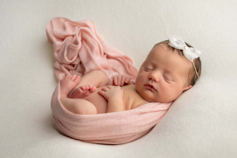Baby girl during newborn photography session, lying on her back with her legs tucked up. Peach wrap around her holding her legs up and white bow in her hair. Taken at Glasgow studio