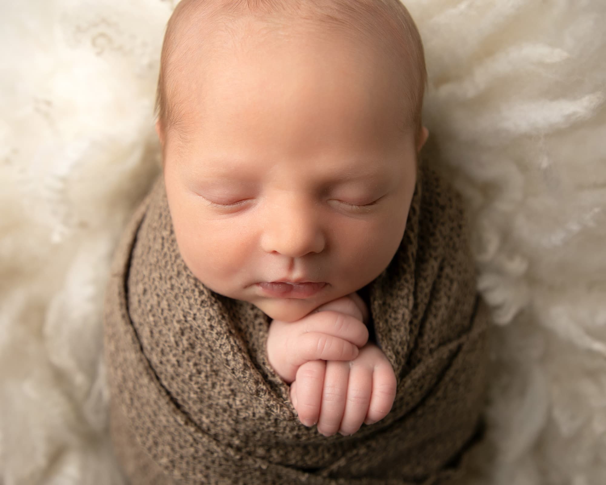 Close up image of baby wrapped in brown fabric with hands below his chin, laying on cream fur. Image provided by Glasgow baby photoshoot