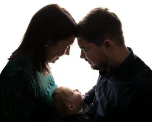 Mum, Dad & Little boy in silhouette image. Mum & dad are facing each other with heads touching. Baby boy is looking up at them. Image taken by Glasgow baby photographer