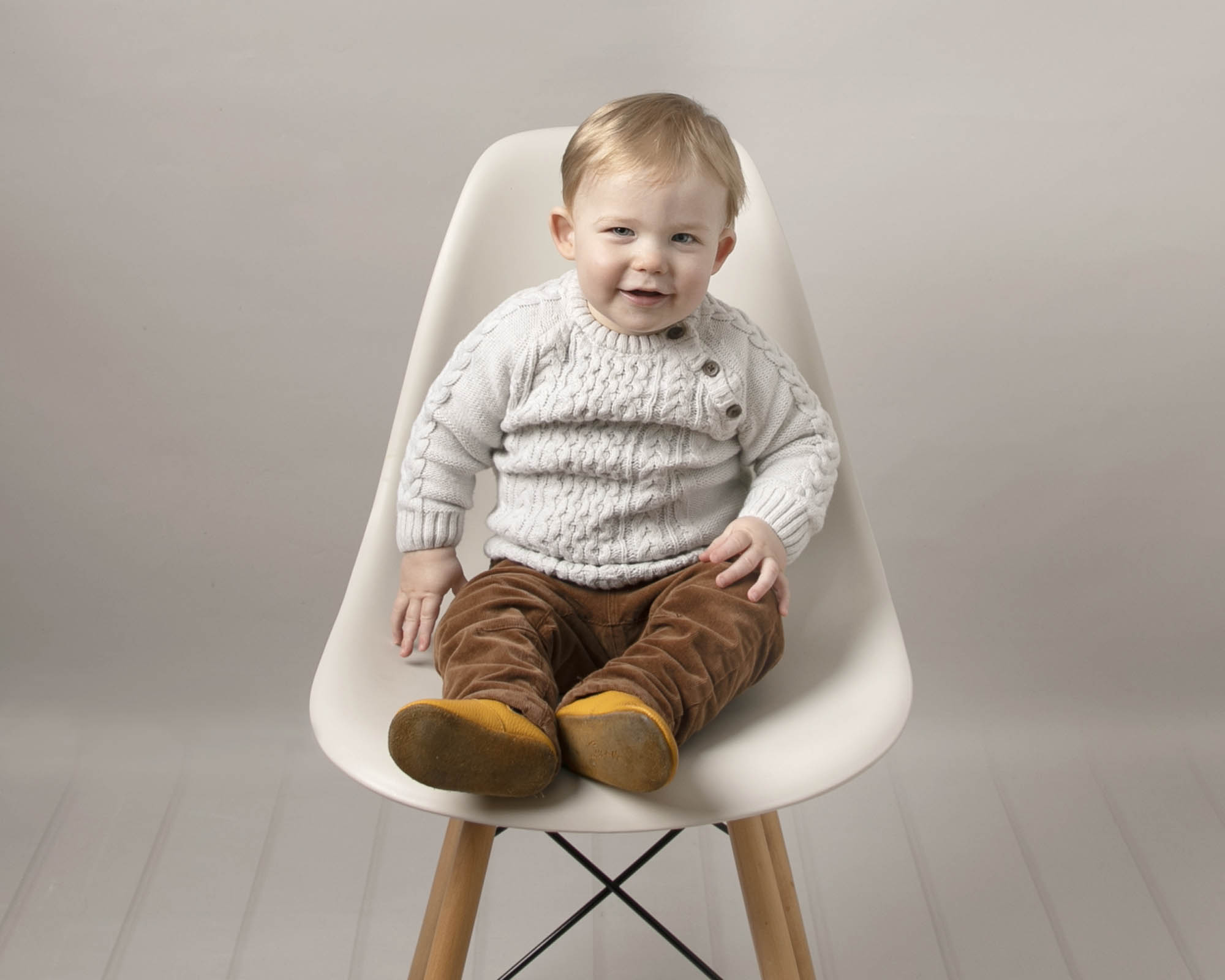 Baby boy sat on a cream hermes chair, wearing brown cord and cream knit jumper. Baby is smiling at his baby photography session with Glasgow photographer