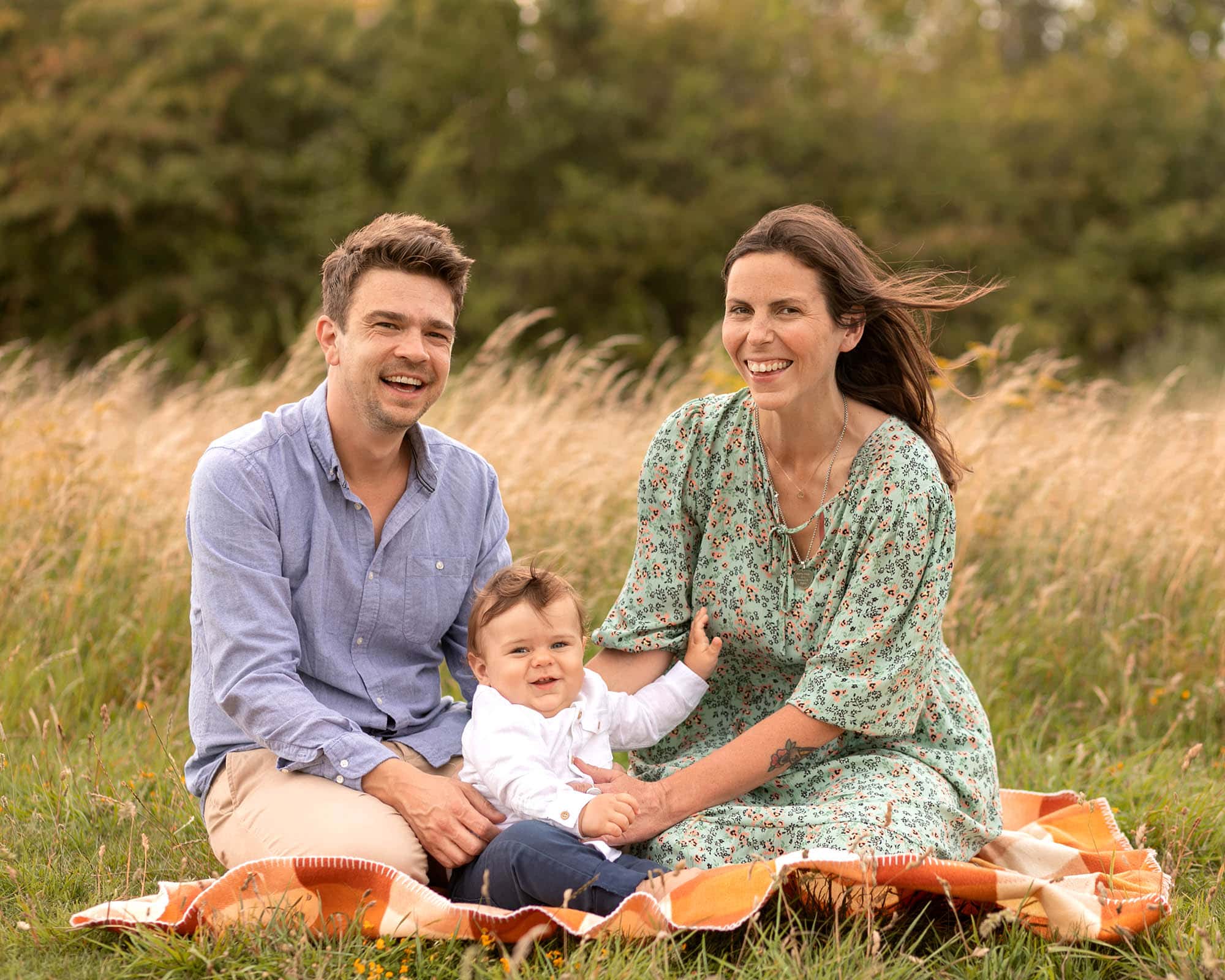 Family sat on orange checked blanket. Female wearing green floral dress. Baby wearing white shirt and blue jeans. Male adult wearing blue shirt and beige chinos. Wind blowing hair and smiling at the camera. Image taken on location in long grassy area in Glasgow by photographer Louise Ferguson