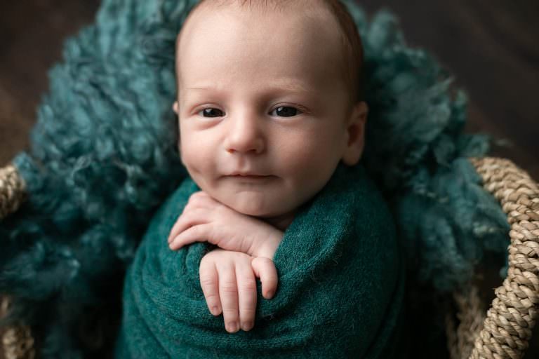 Baby boy in green, in a basket, smiling with his eye open. Part of a gallery from newborn photography shoot in Glasgow