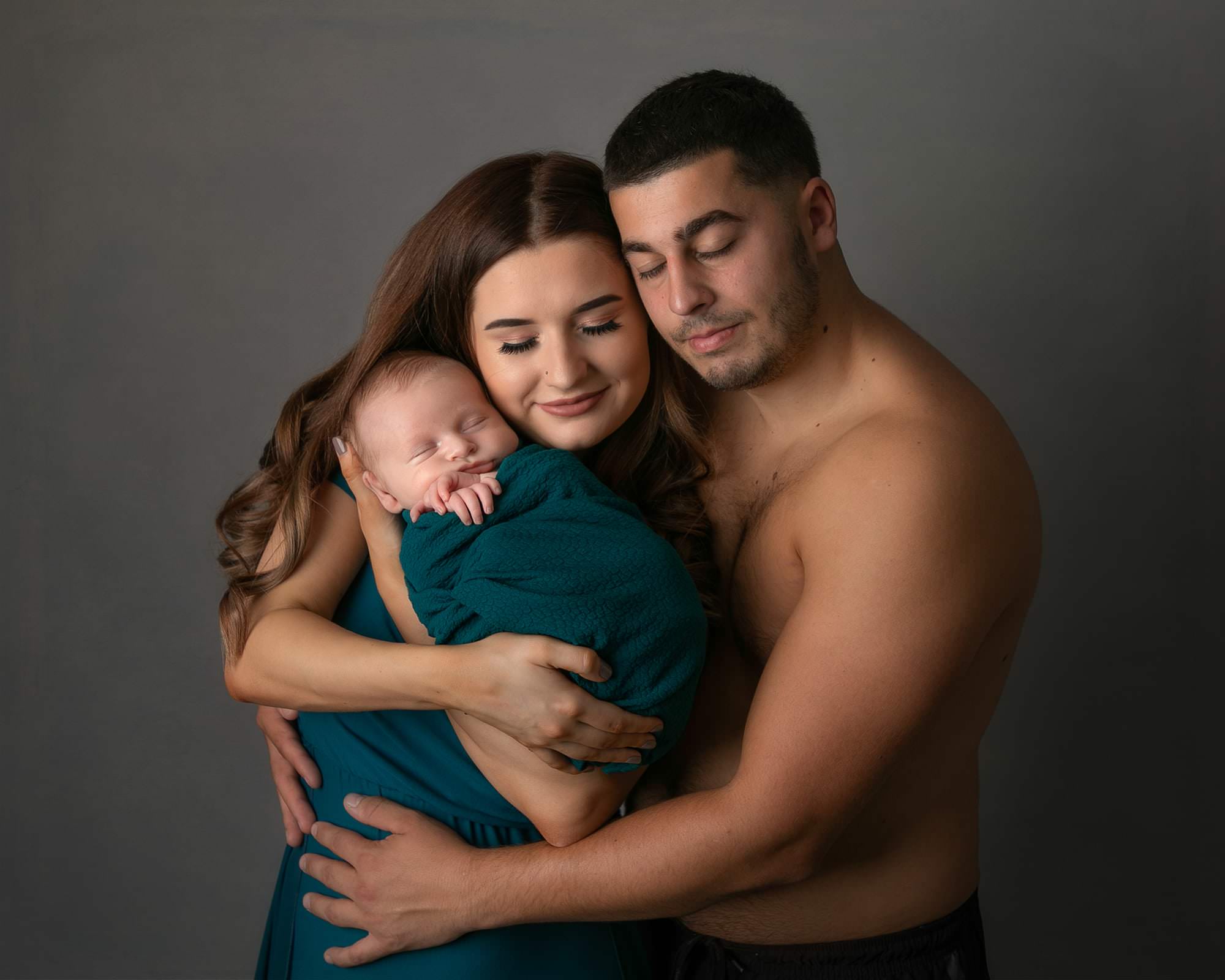 Young male topless, with arm around young girl with long wavy hair. Girl is wearing a green dress holding a baby wrapped in green. Both have their eyes closed. Image taken by Glasgow baby photographer