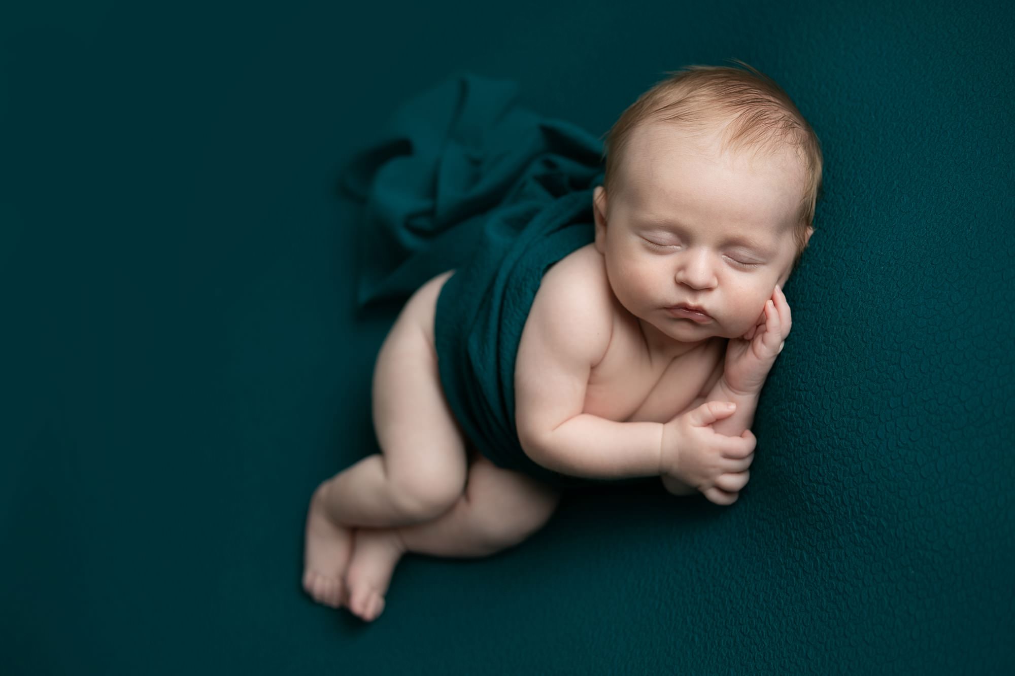 Baby boy lying on his side on a jade green backdrop, with a wrap around his tummy. Image taken by Glasgow photographer during newborn photography session