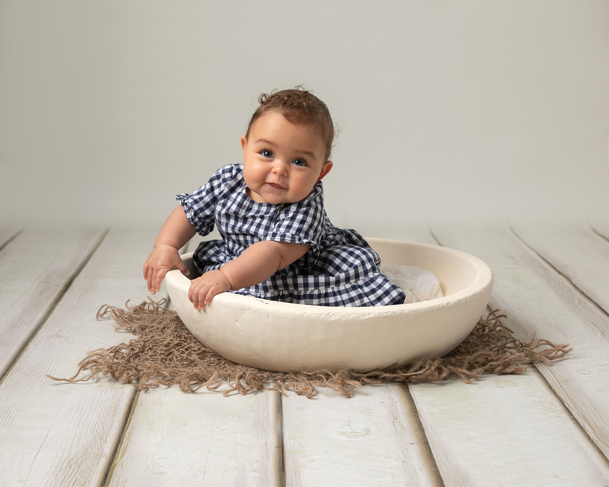 Baby girl in blue & white gingham dress sat in a cream bowl, leaning to the side and looking forward. Baby is having a photoshoot with Glasgow photographer