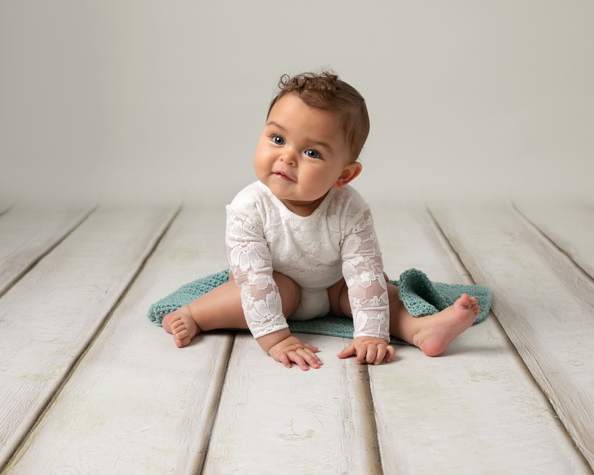 8 month old baby sat on a cream wooden floor on a small blanket wearing a white long sleeve romper. Taken by Glasgow Baby Photographer Louise Ferguson