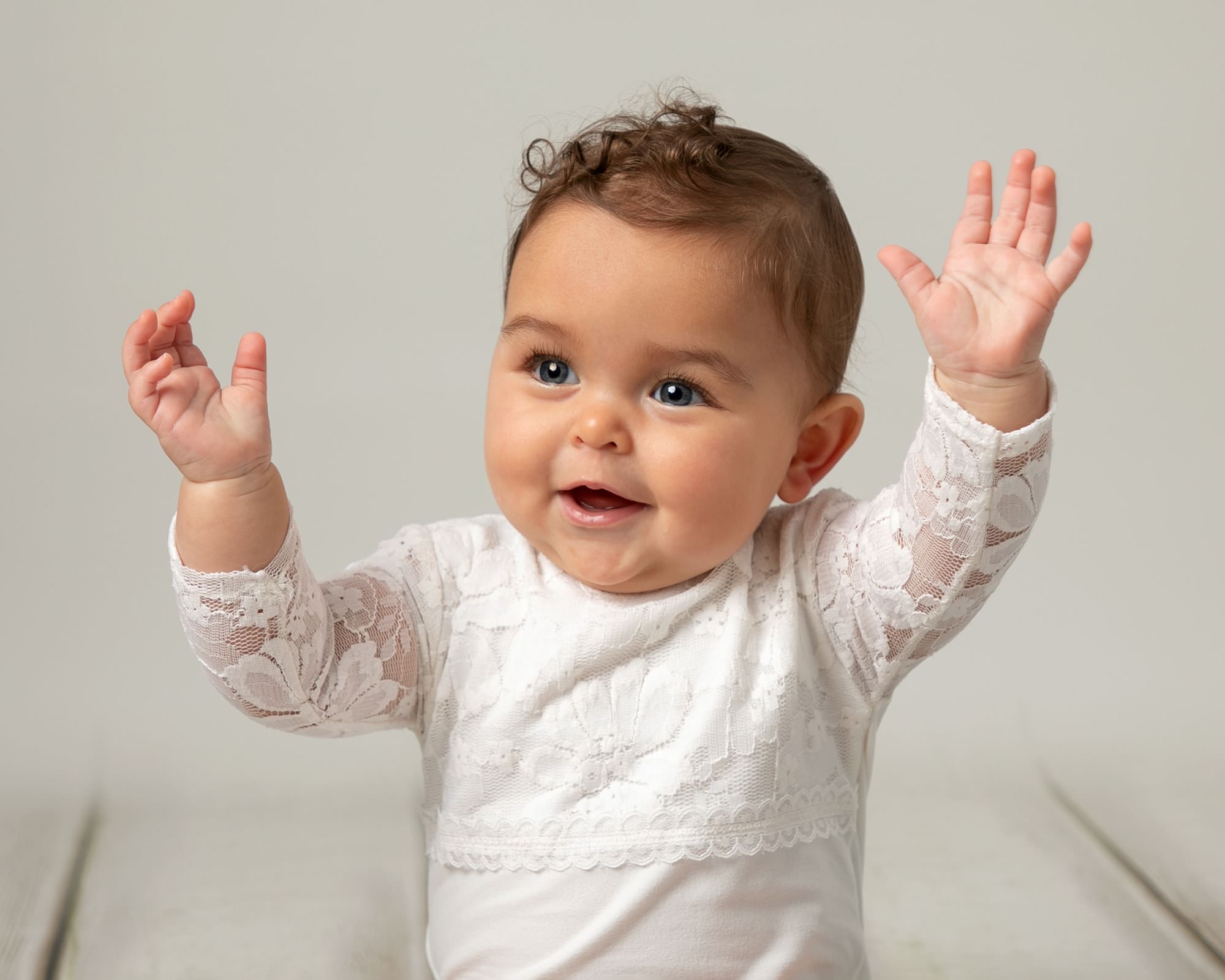 Baby girl with short curls wearing a white romper during a baby photography session in Glasgow. Baby is smiling and holding her hands in the air