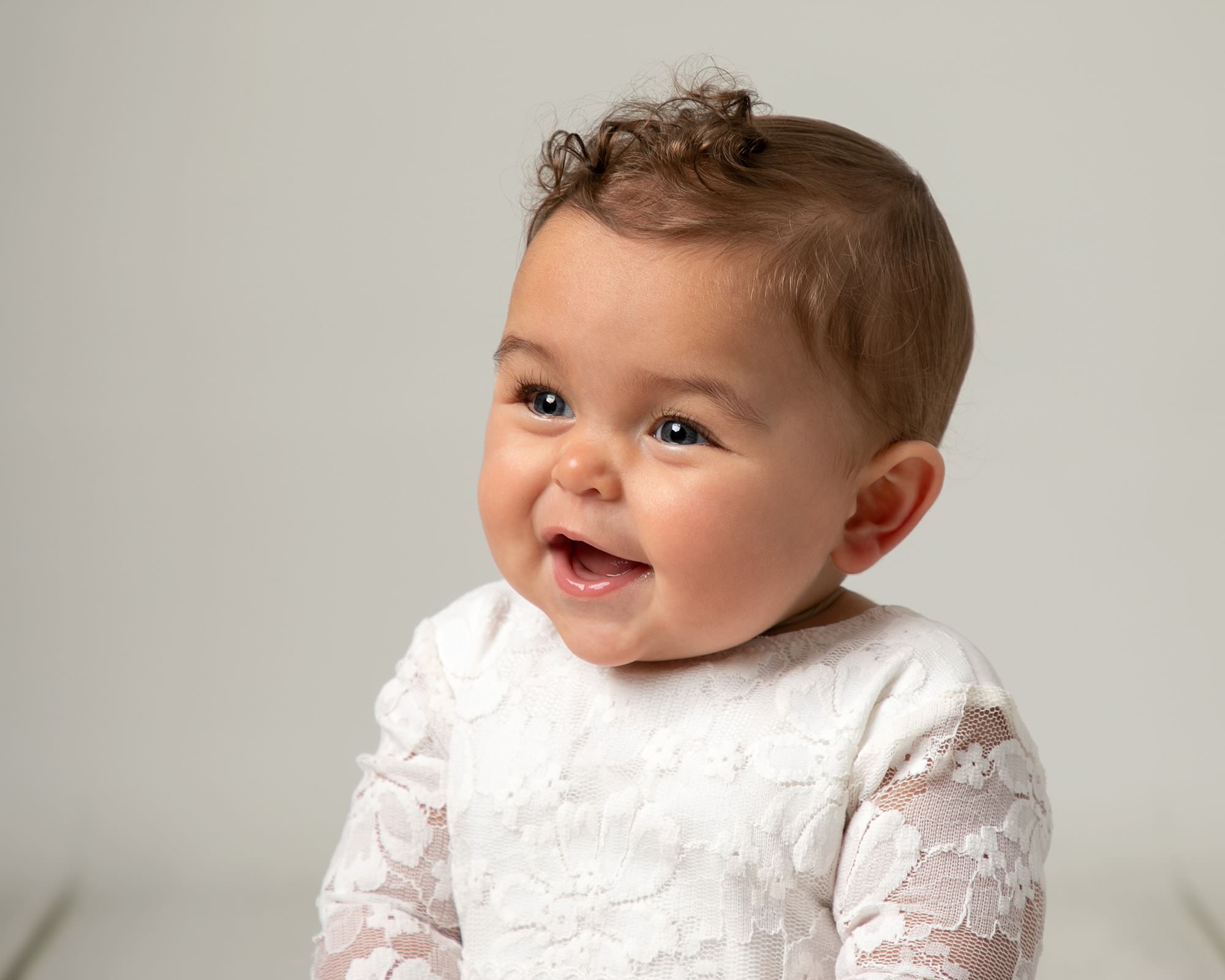 Baby girl with short curls wearing a white romper smiling. Image taken at Glasgow baby photography shoot