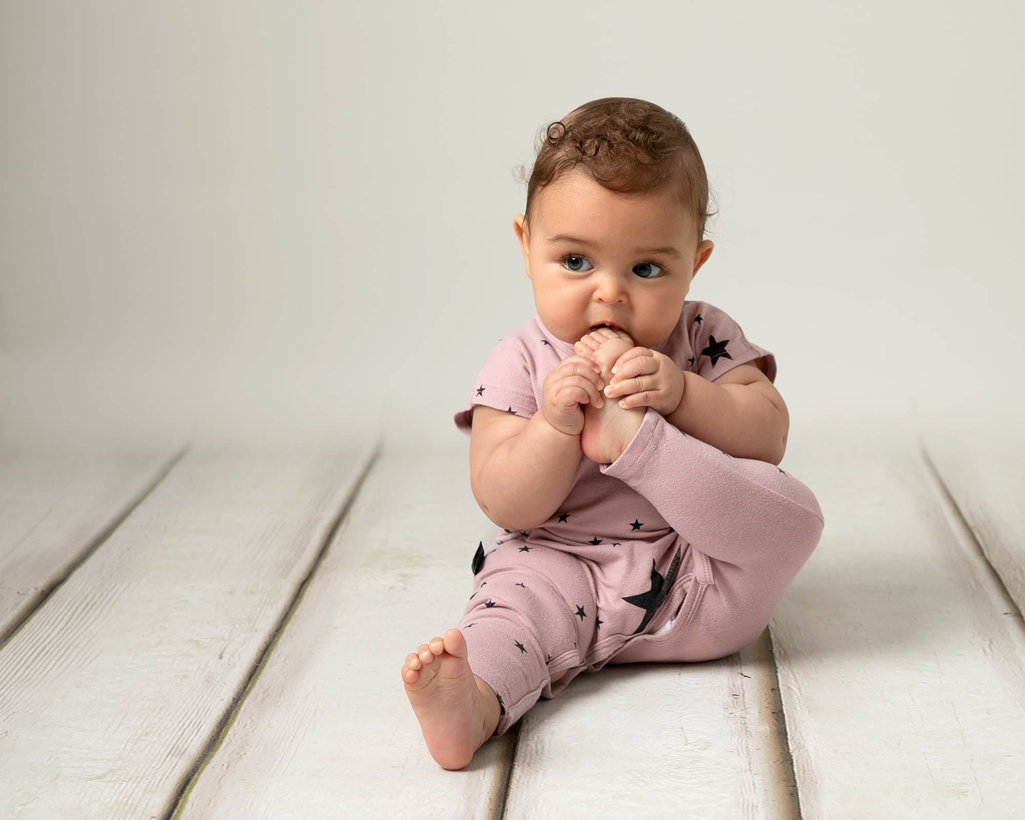 Baby girl with short brown curls wearing a pink romper with blue stars sitting on a cream wood effect backdrop. baby is taking part in a photography session in Glasgow. Toes in hand and in mouth