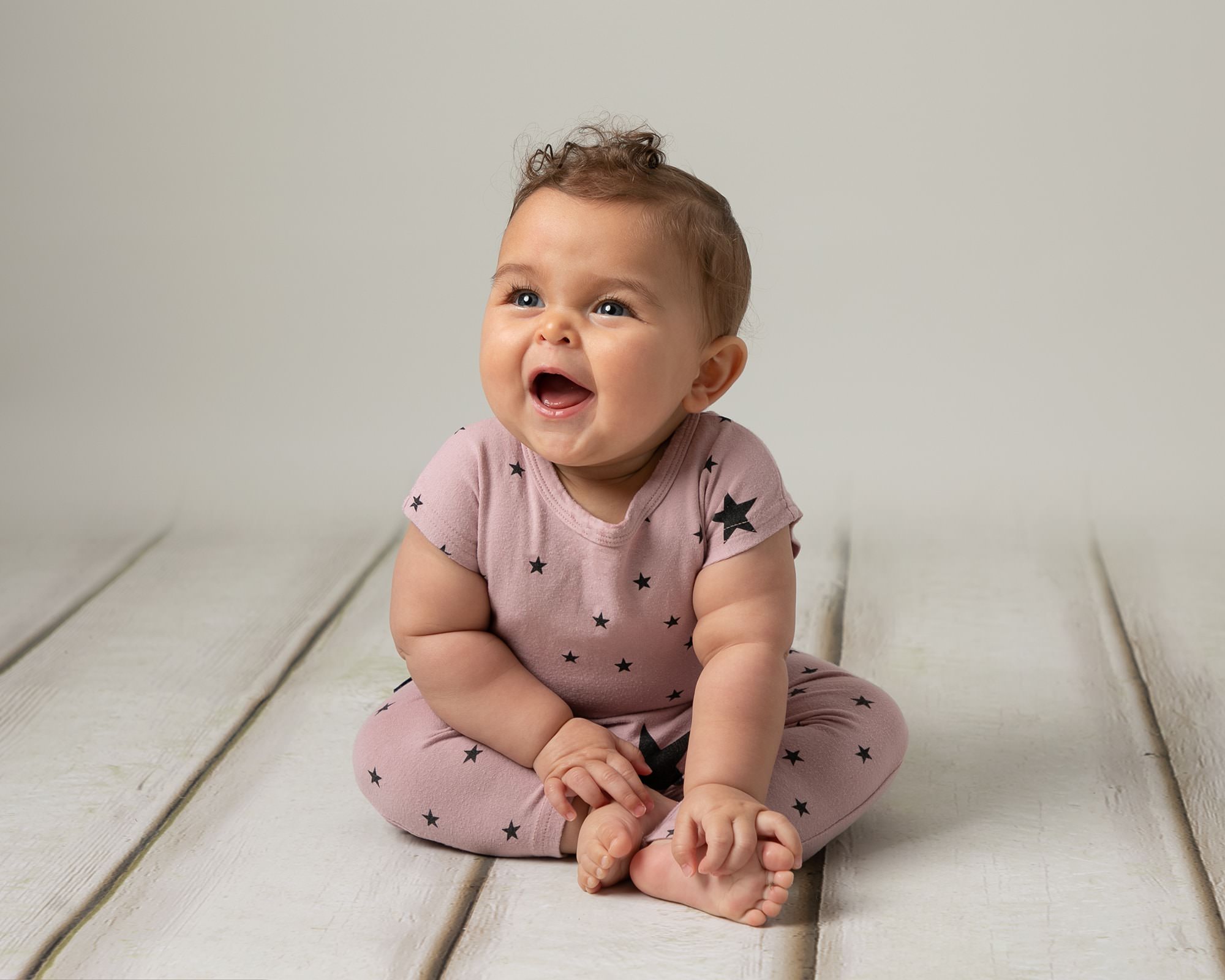 Baby girl smiling at her parent in a baby photoshoot in Glasgow. Girl wears a pink romper with blue stars