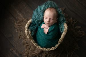 Baby boy wrapped in jade green in a basket on green fur, sat on dark wooden floor. Picture taken by Glasgow babyphotographer