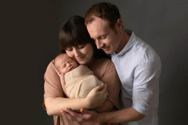 Parents with baby. Mum in pink top, baby wrapped in cream fabric, dad wearing blue shirt. Image part of newborn photography session in Glasgow