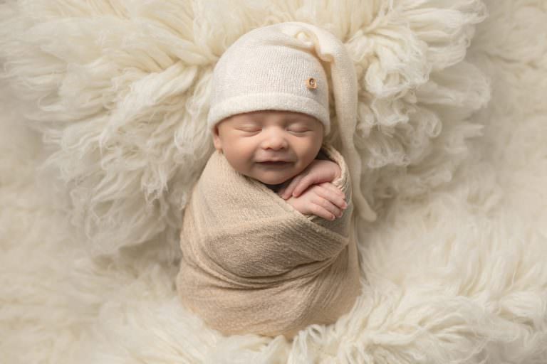 Baby boy swaddled in cream wrap on cream fur flokatti, wearing a cream sleepy hat.