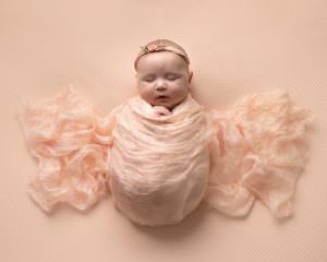 Baby girl on peach blanket swaddled and covered in a peach silk fabric, given the illusion of butterfly wings. Image is taken from above. Image by Glasgow photographer in Newborn photoshoot