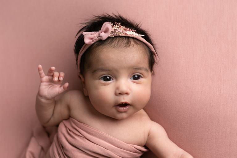 Baby girl on pink blanket with dark hair looking up into the camera. Image taken by Glasgow newborn photographer at photoshoot