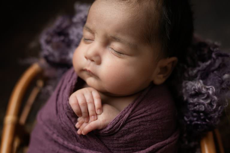 Baby wrapped in purple, close up sleeping with hands crossed. Image part of newborn photography session in Glasgow