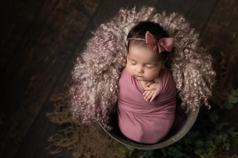 Baby girl wrapped in deep pink fabric wearing pink bow halo, posed in bucket. Image part of newborn photography session in Glasgow
