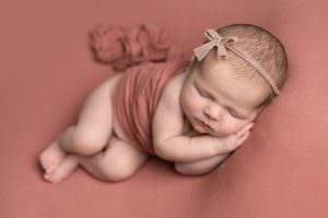 Baby girl on dusky pink backdrop lying on her side, with matching pink fabric around her waist. Baby is wearing a delicate pink fabric halo with a bow. Image part of a newborn photography gallery by Glasgow photographer