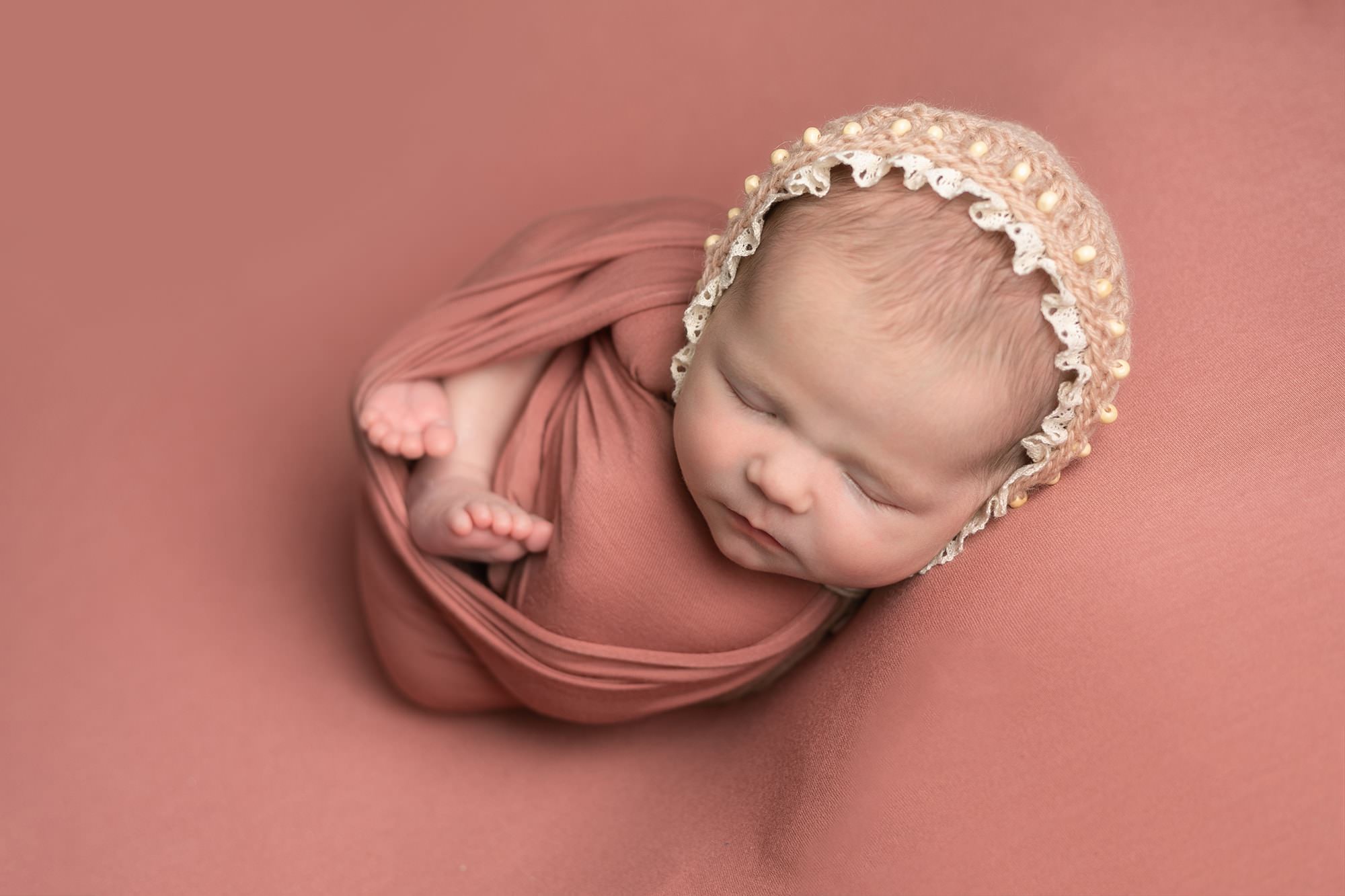 Baby girl on dusky pink backdrop swaddled in wrap of same collier with feet showing. Baby wears a pink bonnet.Image captured during newborn photography session in Glasgow