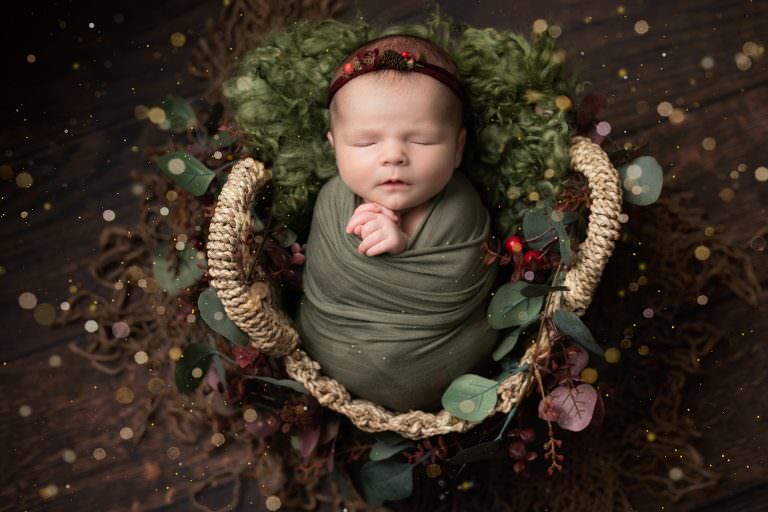 Baby girl swaddled in green wrap, in a wicker basket on a dark wood floor. Baby is resting against a green woven blanket with festive greenery wrapped around the basket. Image part of a newborn photography session in Glasgow, with festive styling.