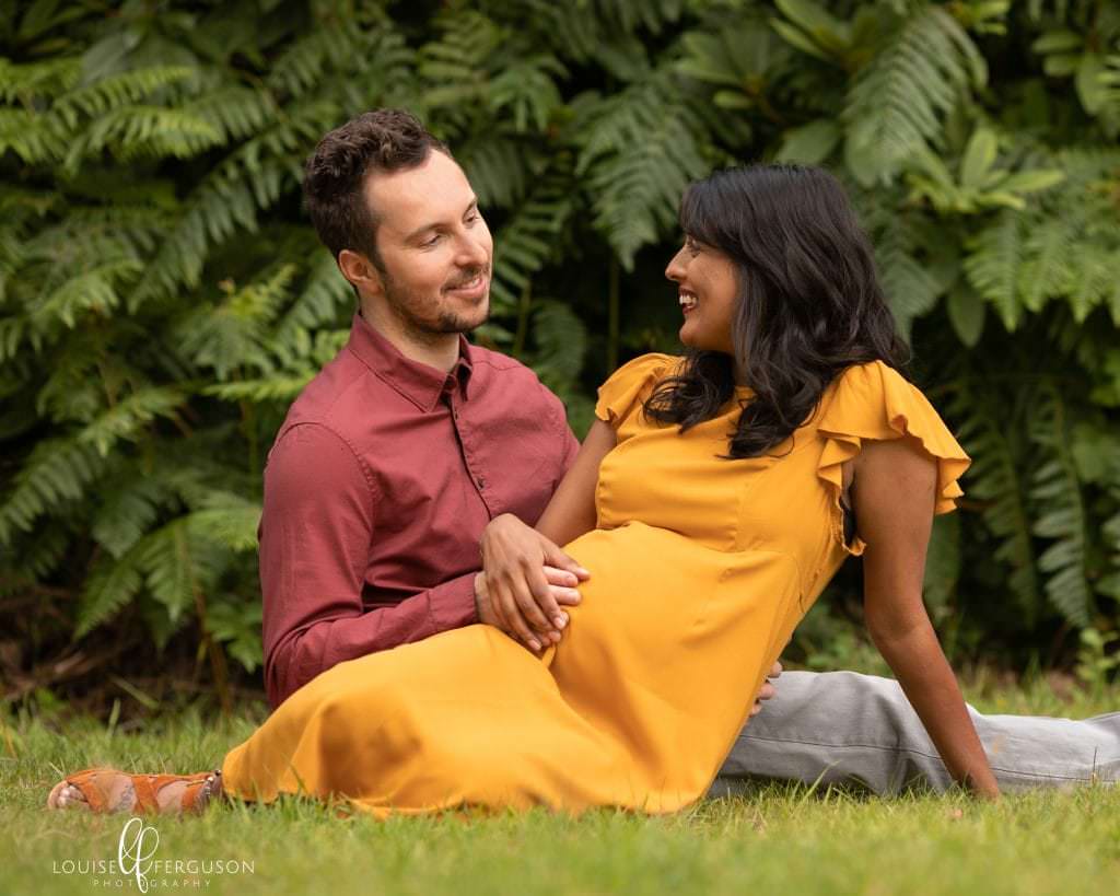 Couple sat outdoors, female wearing a ochre coloured dress, male wearing a rust coloured shirt and beige chinos. Couple are sat on grass with tall ferns behind. Female is pregnant. Image taken during pregnancy photoshoot in a Glasgow beauty spot