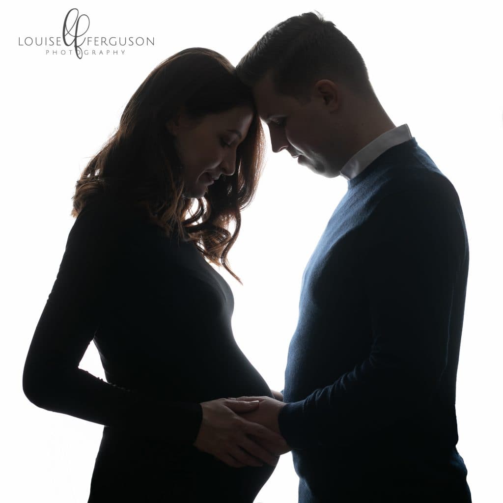 Male & Female wearing dark clothing facing each other, heads touching looking down at baby bum. Image is a silhouette as part of a maternity photography session in Glasgow