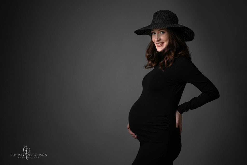 Pregnant female with dark hair wearing a black polo neck, tight fitting dress and a black hat. Part of a gallery of images from a maternity photography in Glasgow studio