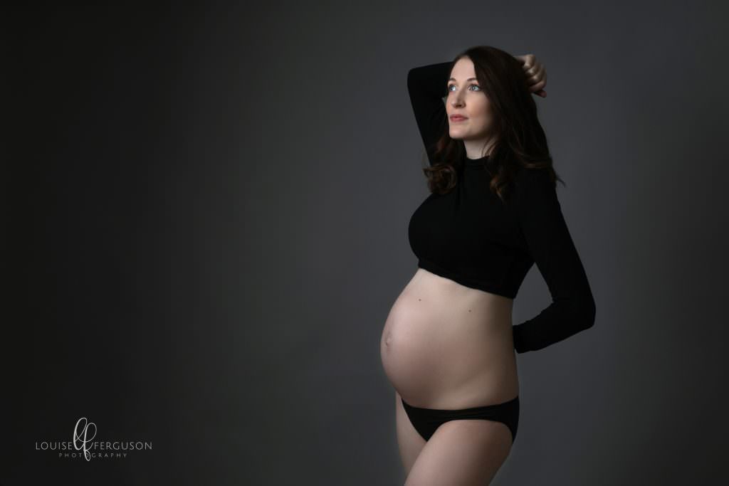 Pregnant female at maternity photography session. Wears a cropped black polo neck and black pants. Standing facing light source at Glasgow studio