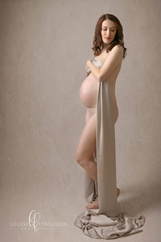 Naked pregnant female, covering her breasts with a satin fabric, posing for maternity photography at Glasgow studio