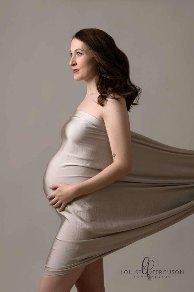 Pregnant female facing left profile view with satin fabric on cream wrapped around her bump. Image taken by Glasgow photographer during maternity photography session
