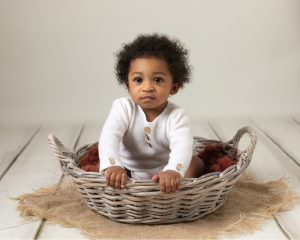 Baby boy wearing white rompers sat in a cream bowl on brown fluff, leaning forward looking at the camera. Image taken by award winning baby photography in Glasgow