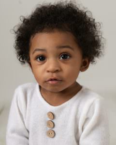 Baby boy wearing cream romper. Boy has brown afro hair and is looking directly at the camera. Photoshoot for older baby