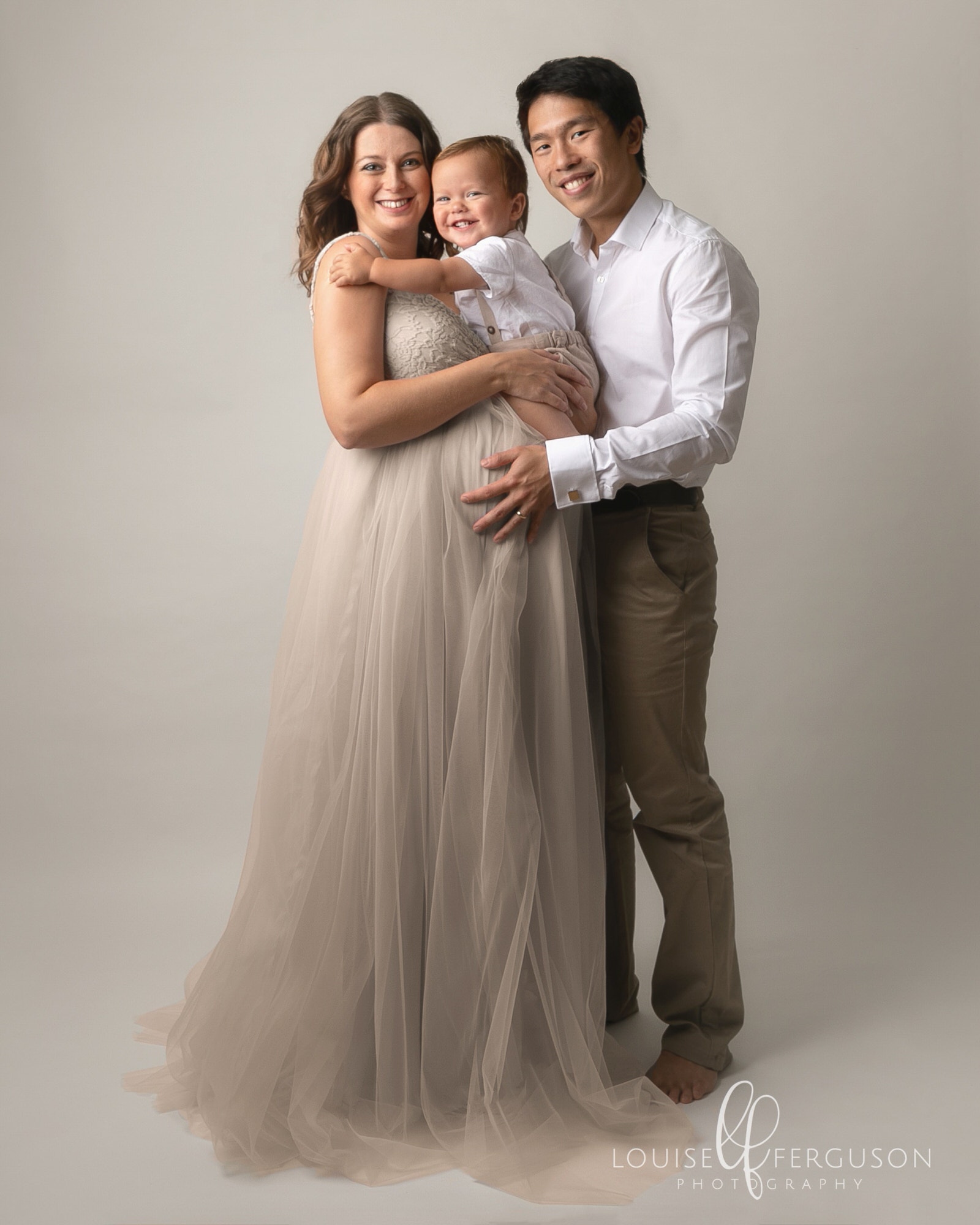 Image taken by Maternity Photographer in Glasgow. Male & female with male toddler. Female is pregnant and wearing a cream gown with tulle skirt, She is holding toddler on her bump and male has his arms around her. Male wears a white shirt & chinos
