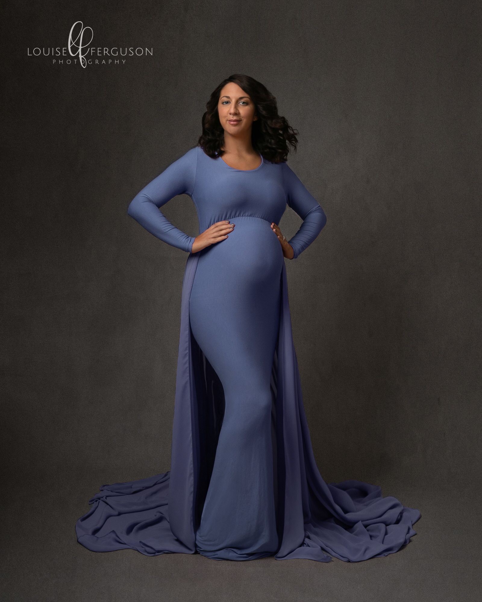 Pregnant female with long dark hair, wearing a violet blue gown, facing forward with her hand on her hips. Full length image taken on a grey backdrop by Glasgow Maternity Photographer