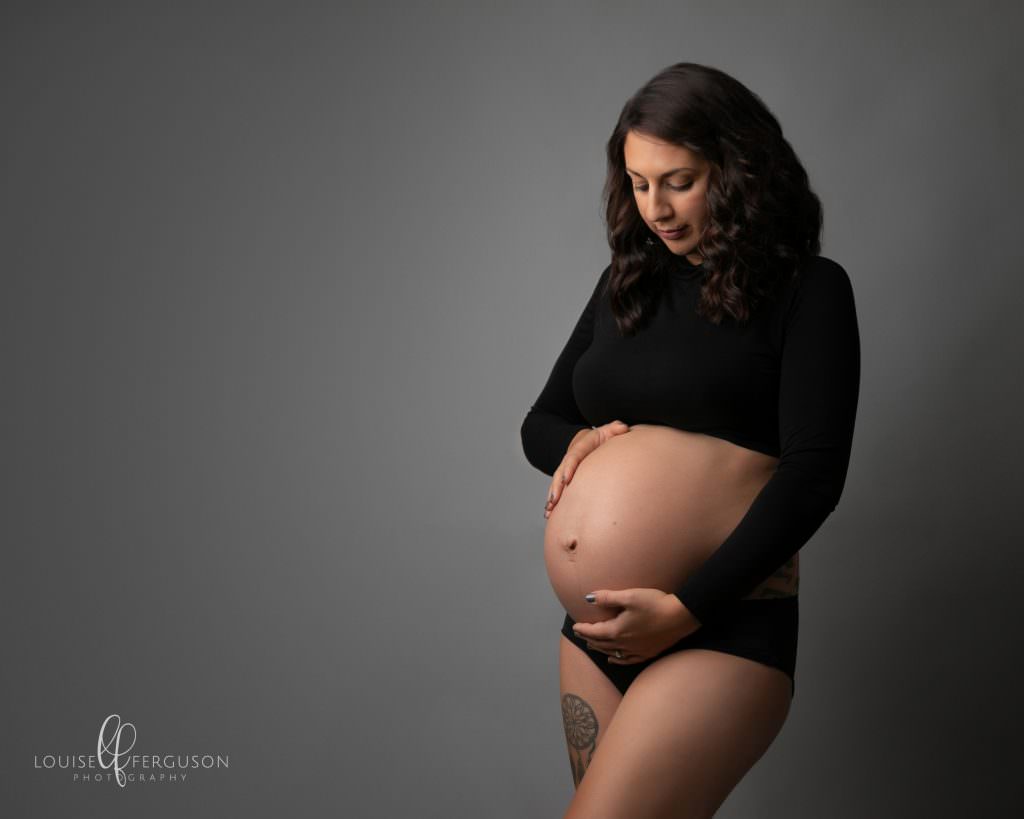 Dark haired female in shot taken at glasgow studio. Looking down at baby bum and smiling. Female wears a black maternity top for her maternity photography session