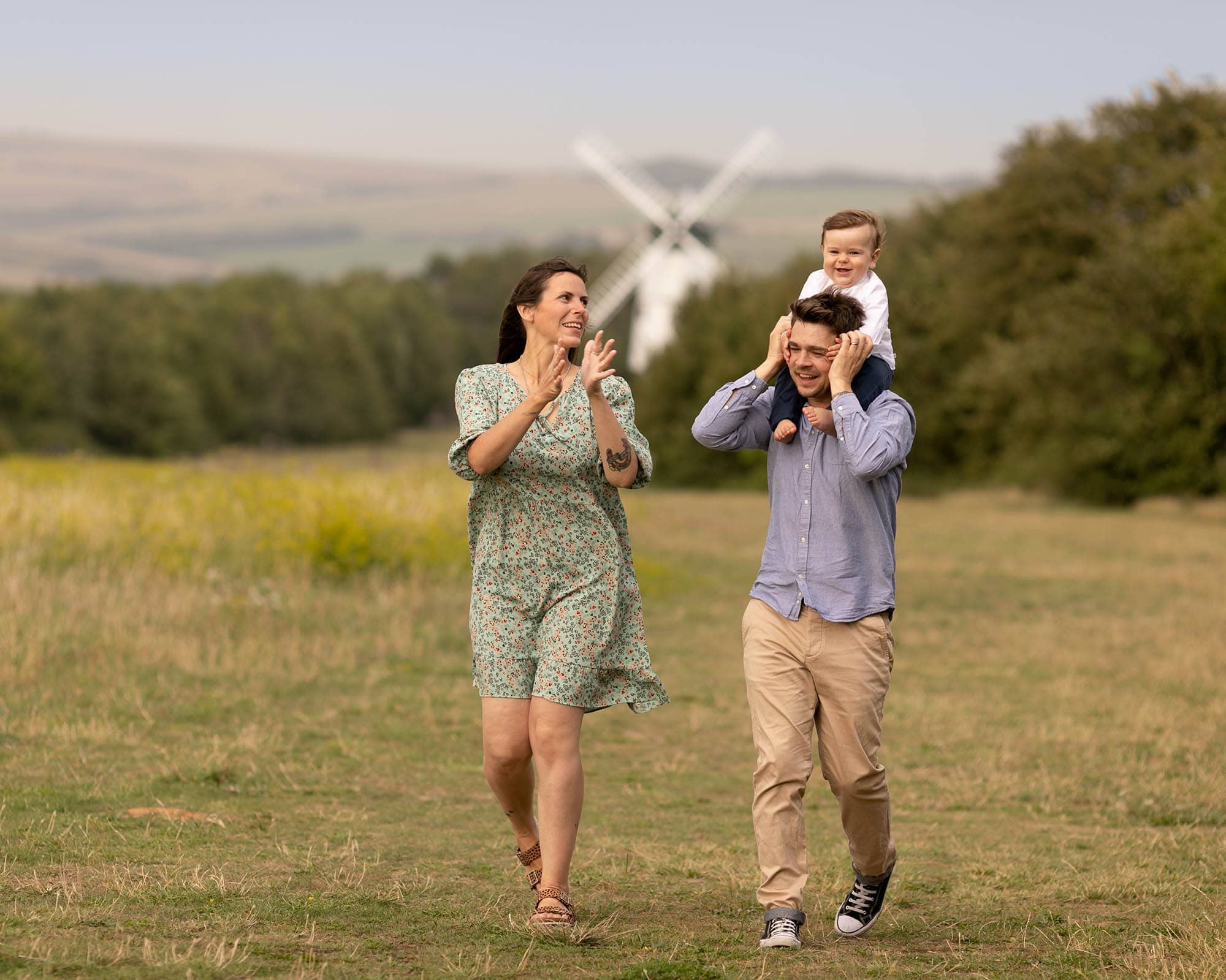 Couple walking in the countryside. dad has baby pn his shoulders. A windmill can be seen in the background. Image taken during a family photography session in Glasgow