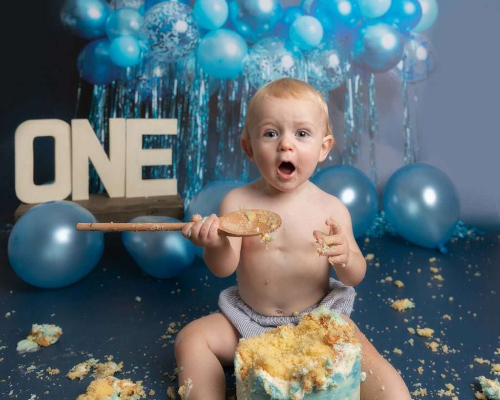 Baby boy sat with a 1st birthday cake and holding a wooden spoon. Boy sits on a blue backdrop with balloons at Glasgow studio during his cakesmash photoshoot