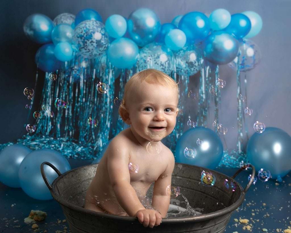 Baby boy in tin bath smiling, blue backdrop with balloons as he celebrates his 1st birthday with a cakesmash photoshoot at Glasgow Studio