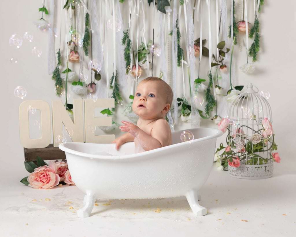Baby girl in white roll top bath in front of pink flower garland as part of 1st birthday cakesmash photoshoot in Glasgow