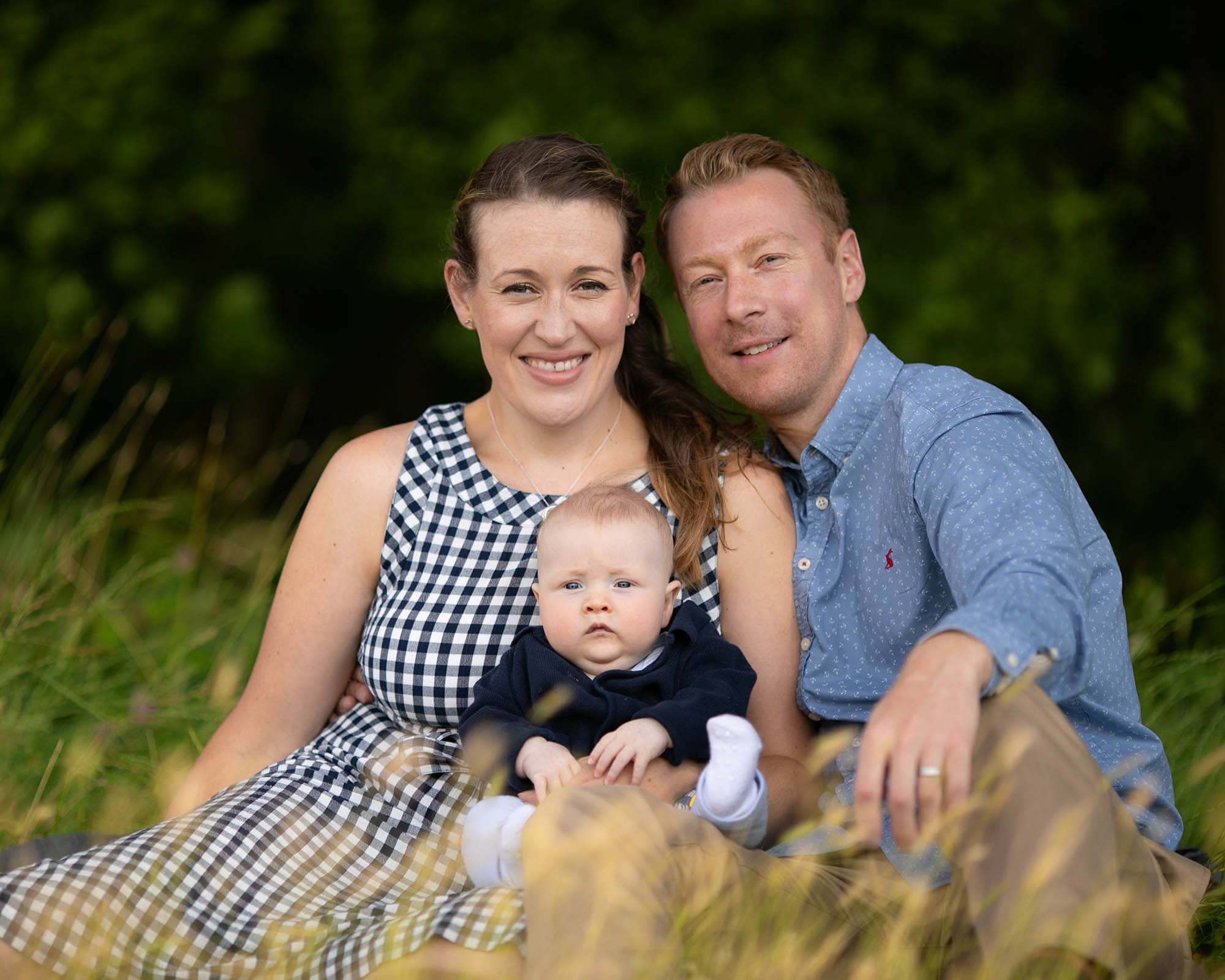 Family taking part in a photography session in Glasgow. Parents all wearing blue and baby on knee, sat in long grass