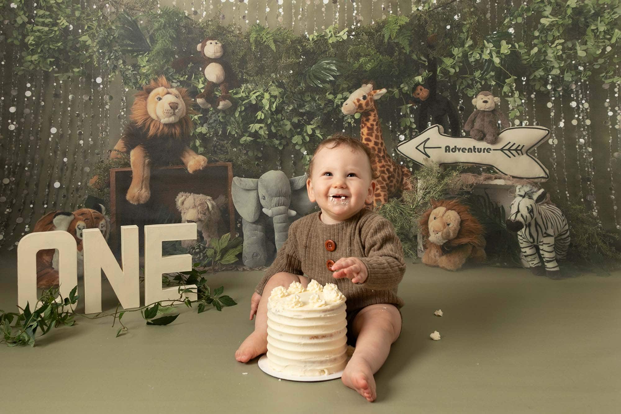 Baby boy sat with cream 1st birthday cake, in front of a jungle scene backdrop, smiling at the camera. Image part of a cakesmash & splash.