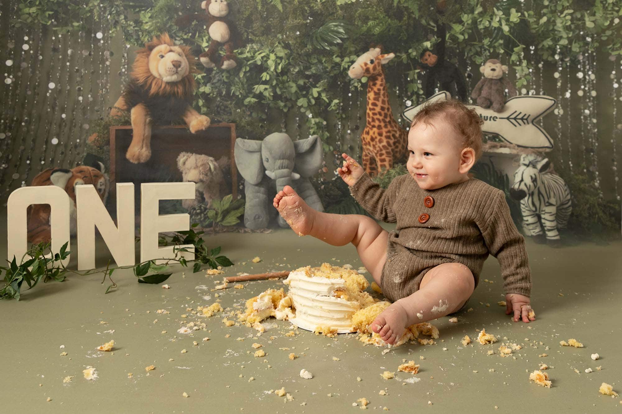 Baby boy in brown romper sat on jungle backdrop, kicking his 1st birthday cake. image taken at cakesmash photoshoot in Glasgow