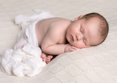 Baby boy on cream embossed blanket lay on tummy with white fabric covering him, Image taken during newborn baby photoshoot in Glasgow