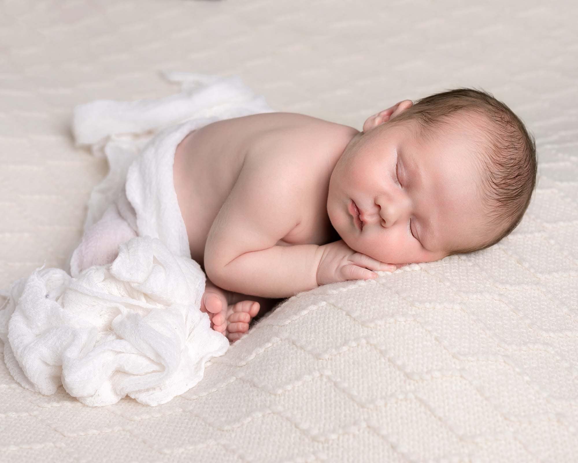 Baby boy on cream embossed blanket lay on tummy with white fabric covering him, Image taken durin baby photoshoot in Glasgow