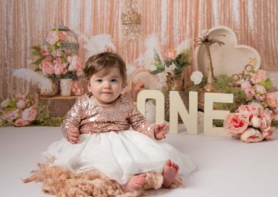 Baby girl in sparkly peach dress with white tulle, sat on the floor smiling. Vintage backdrop in peach tones with cream one letters.