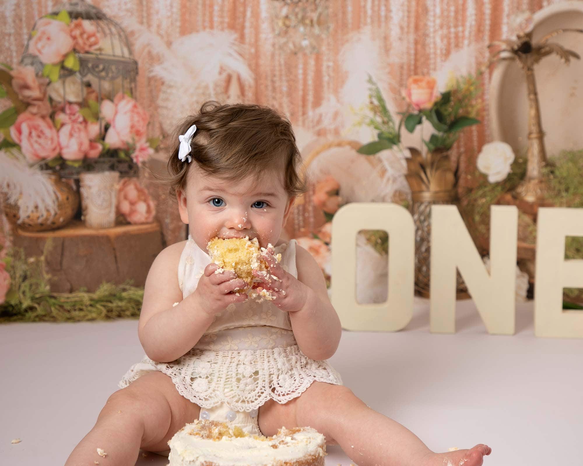 Baby girl in cream romper eating cake. Image at her 1st birthday photoshoot in Glasgow. Vintage backdrop in peach tones with cream one letters.