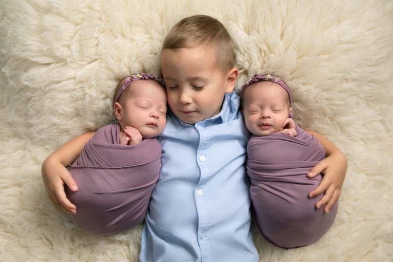 Twin girls wrapped in purple wraps during newborn photography session in Glasgow. Girls are lying on a cream fur with their big brother who wears a blue shirt