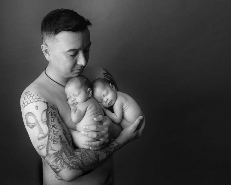Black & white image of dad holding newborn twin babies against his bare chest. Image taken during newborn baby photoshoot in Glasgow