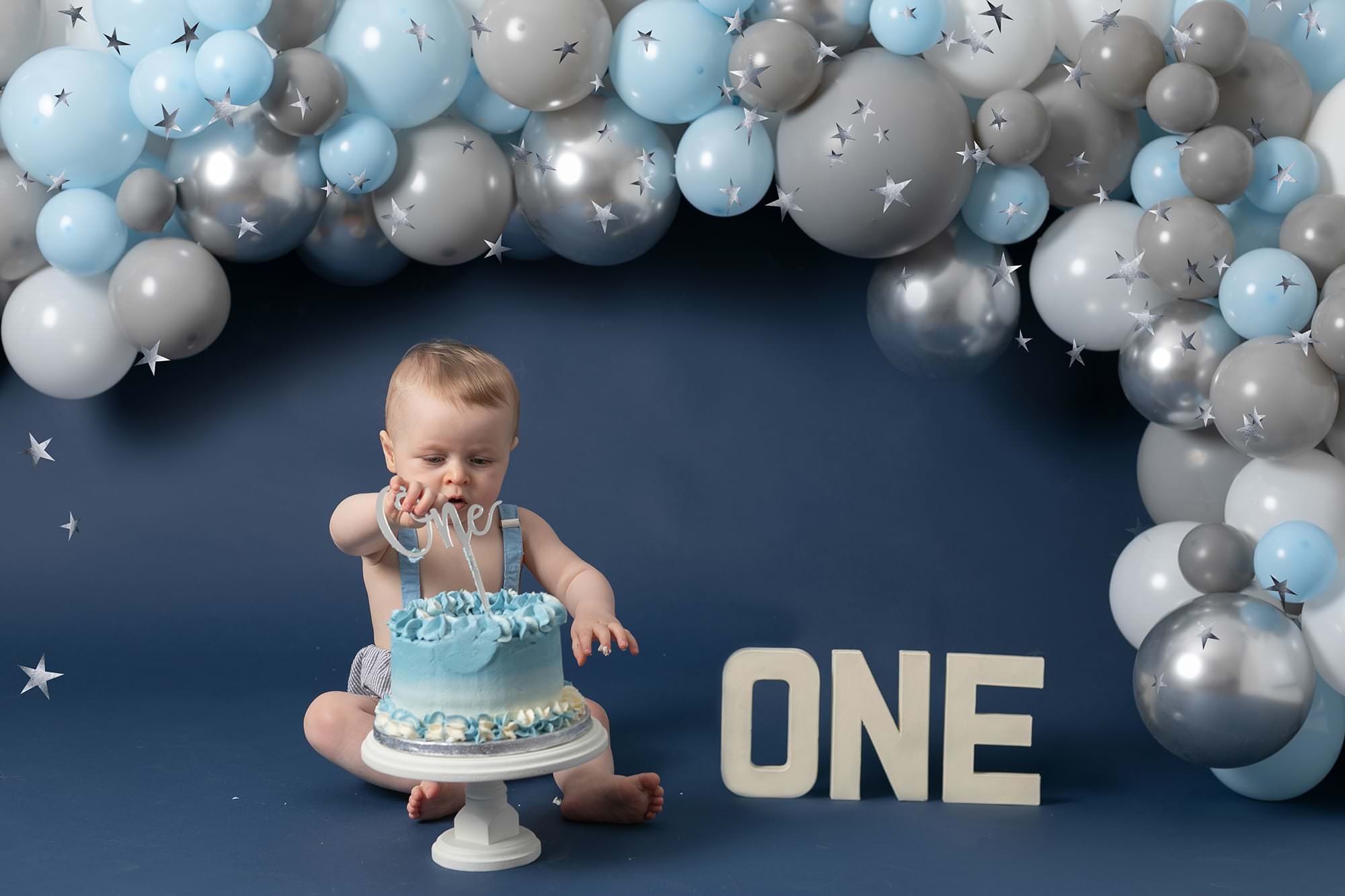 Baby boy on blue backdrop at his Birthday cakesmash photoshoot. Baby is sat behind a cake on a cake stand with a blue, grey & white balloon garland behind him and ONE letters in cream