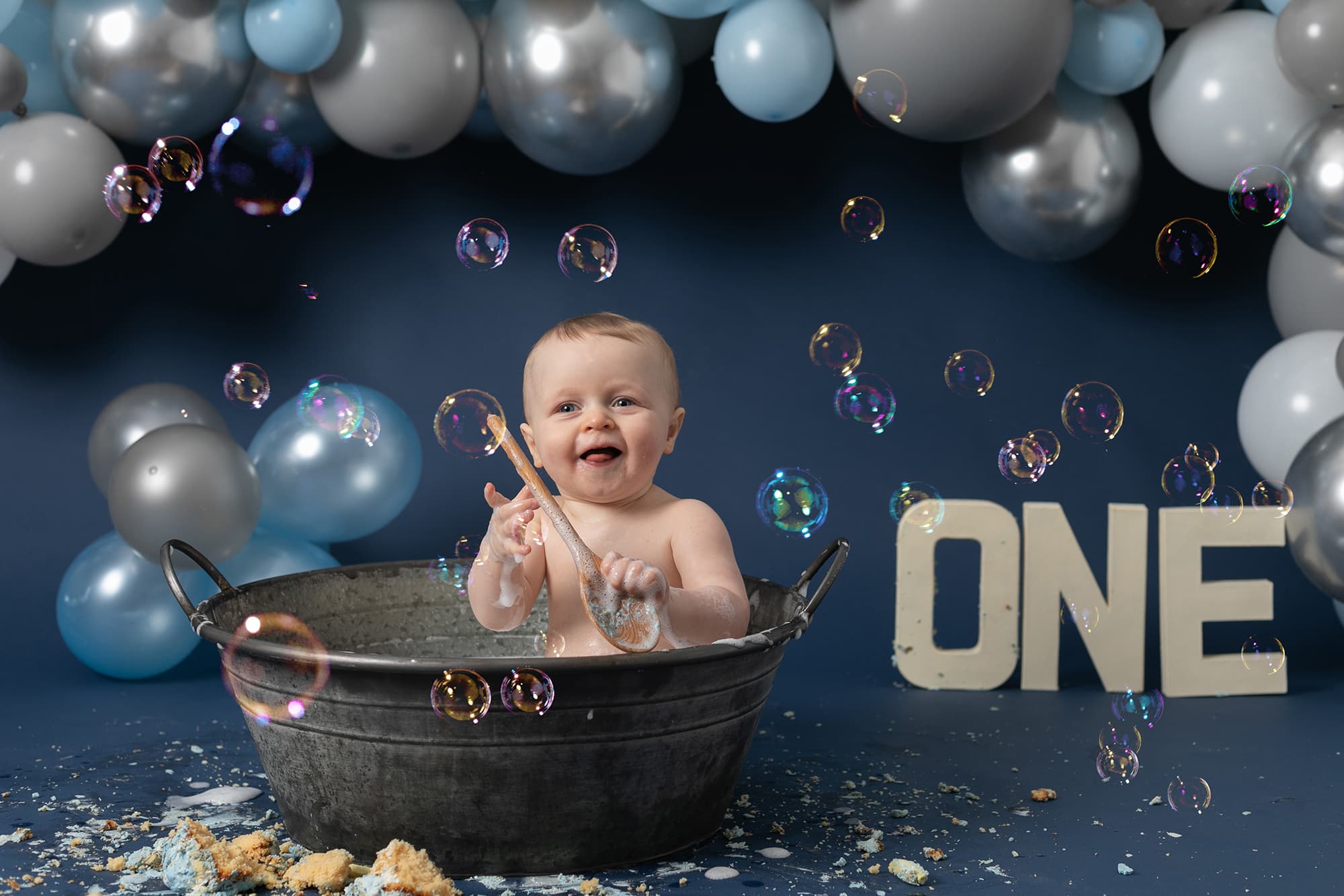 Baby boy on blue backdrop at his 1st Birthday Cakesmash Photoshoot in Glasgow. Baby is sat in a tin bath with a blue, grey & white balloon garland behind him and ONE letters in cream